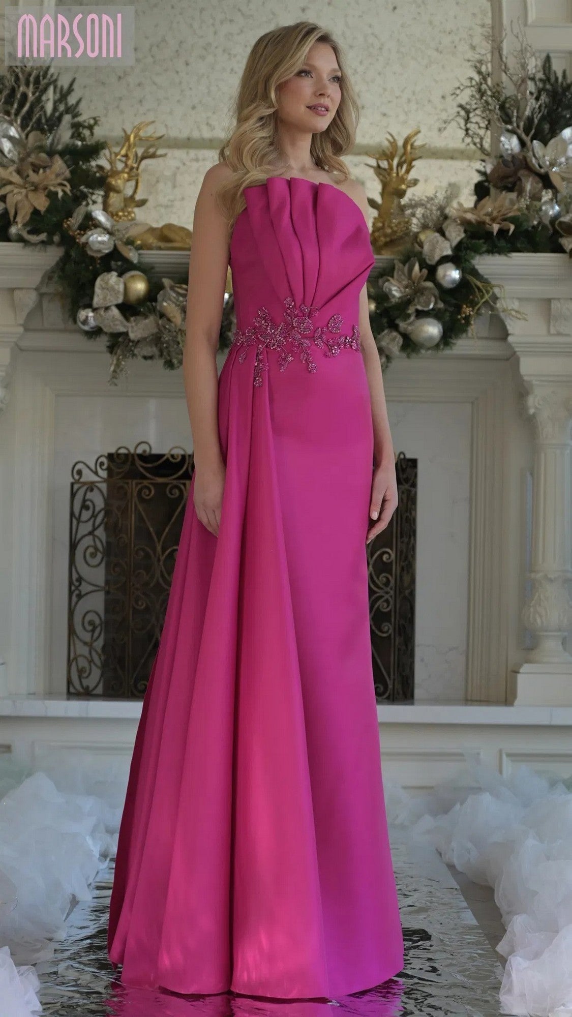 A woman stands indoors wearing the Long Formal Dress: Marsoni MV1356, featuring a vibrant pink color, pleated detailing, and floral embellishments at the waist. Decorative greenery and ornaments appear in the background.